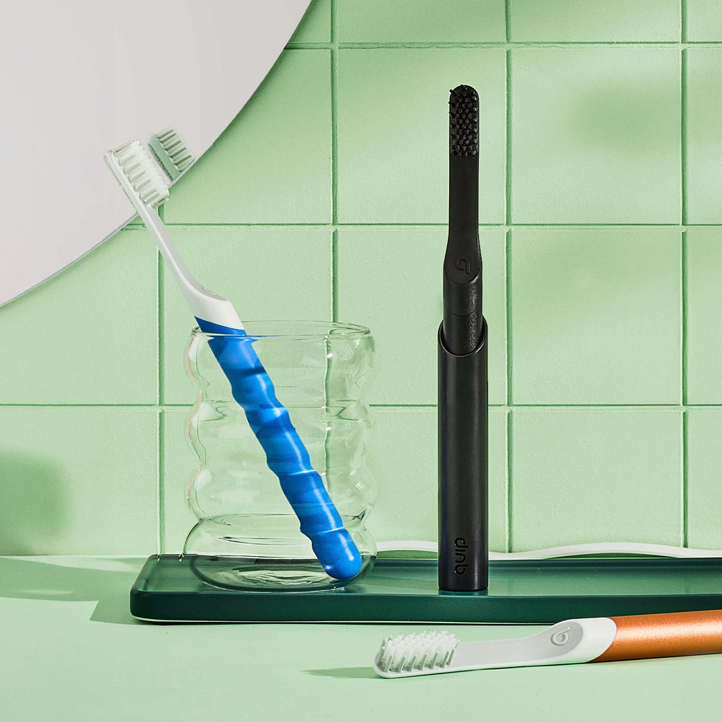 Bathroom counter scene with a black Sonic toothbrush standing upright in its holder beside a clear glass containing a blue-handled toothbrush, with another toothbrush lying in the foreground against a light green tiled wall.