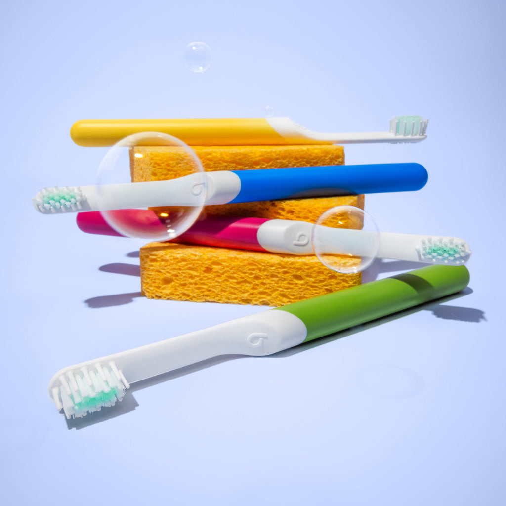 Four colorful quip Kids Sonic toothbrushes—yellow, blue, pink, and green—arranged around two orange sponges with floating soap bubbles against a light purple background.