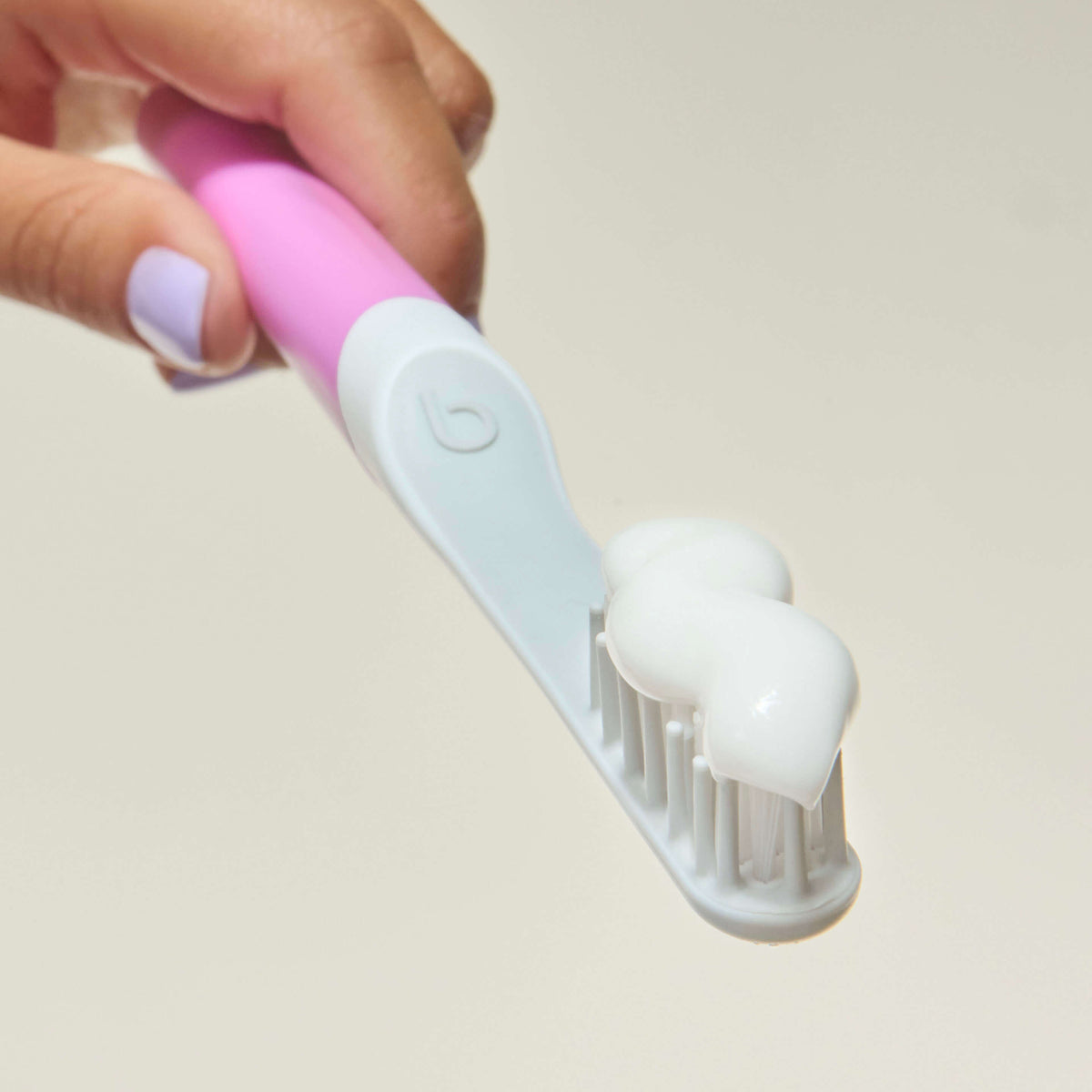 Close-up of a pink and light blue toothbrush head with soft white bristles, topped with a swirl of white toothpaste, held by a hand with lavender-painted nails.