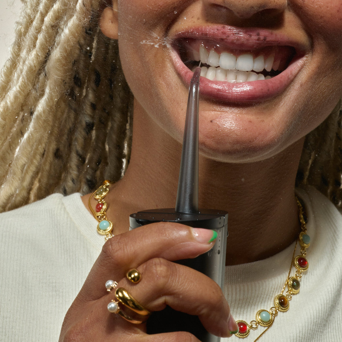 Close-up of a smiling person using a handheld water flosser to clean between their upper front teeth, with water droplets visible and gold jewelry on their hand and neck.